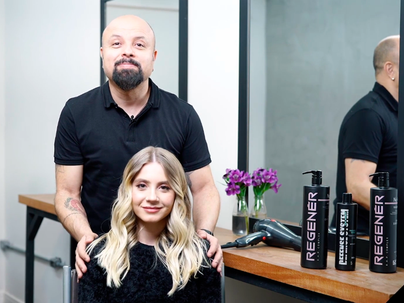 A imagem mostra uma mulher loira usando camiseta preta e blazer preto sentada no centro. Atrás dela está um homem em pé com cabelos escuros e uma camisa preta.